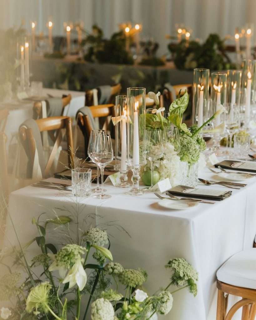 Festlich gedeckte Hochzeitstafel mit weißen Tischdecken, Glaszylindern mit Kerzen, grüner Floristik und eleganter Tischdekoration